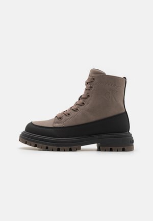 Bottines mi-hautes en daim marron avec accents en caoutchouc noir, bout rond et semelle texturée. Présente des œillets de lacet et un détail de logo en relief.
