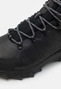 Botte de randonnée noire en cuir texturé, semelle durable, languette en mesh et lacets ronds gris passés dans des œillets métalliques.