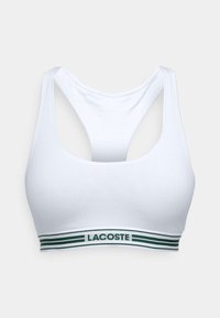Balta sporta krūšturis ar ovālu dekoltē un sporta aizmuguri. Iezīmēta ar zaļi un baltu svītru joslu ar "LACOSTE" logo. Mīksta auduma.