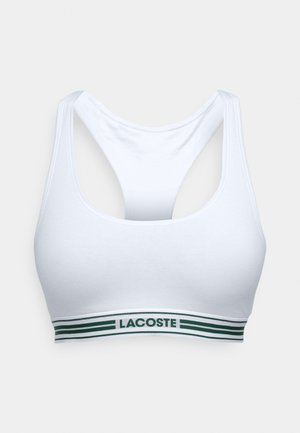 Balta sporta krūšturis ar ovālu dekoltē un sporta aizmuguri. Iezīmēta ar zaļi un baltu svītru joslu ar "LACOSTE" logo. Mīksta auduma.