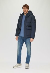Marineblaues Steppjacke mit Kapuze, Reißverschluss und mehreren Taschen, über einem hellblauen Pullover getragen. Kombiniert mit hellblauen Jeans und grauen Sneakern.