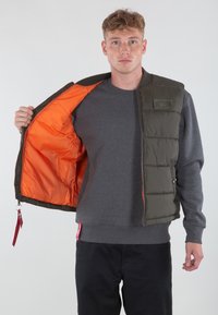 Gilet imbottito verde oliva con foderato arancione, presenta una chiusura con zip, tasche laterali e una toppa con logo. Indossato sopra una felpa grigia.