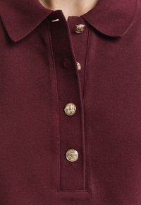 Close-up of a maroon polo shirt with a rounded collar and four gold buttons down the placket.