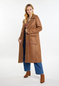 Brun kunstlæderfrakke med en tekstureret shearlingkrave og lomme, udstyret med sølvknapper og en lang pasform, båret over blå denim.