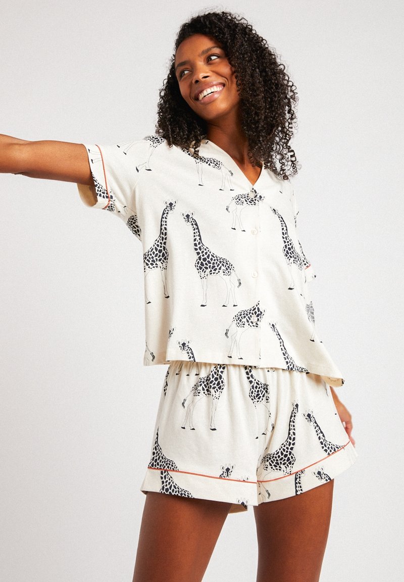 Conjunto de pijama ligero con fondo color crema y estampado de jirafas negras. Camiseta de manga corta con cuello y pantalones cortos con cintura elástica.