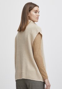 Beige bordás ujjatlan pulóver magas nyakkivágással, világosbarna hosszú ujjú felsőre húzva. Puha textúra és laza szabás.