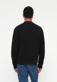Zwarte gebreide trui met een ronde halslijn en geribde manchetten, gedragen met blauwe denim jeans. De trui heeft een wafelpatroon.