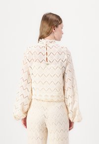 Top in pizzo crema con maniche lunghe e a sbuffo, colletto alto e chiusura con bottoni sul retro. Presenta pattern zigzag e geometrici. Texture liscia.