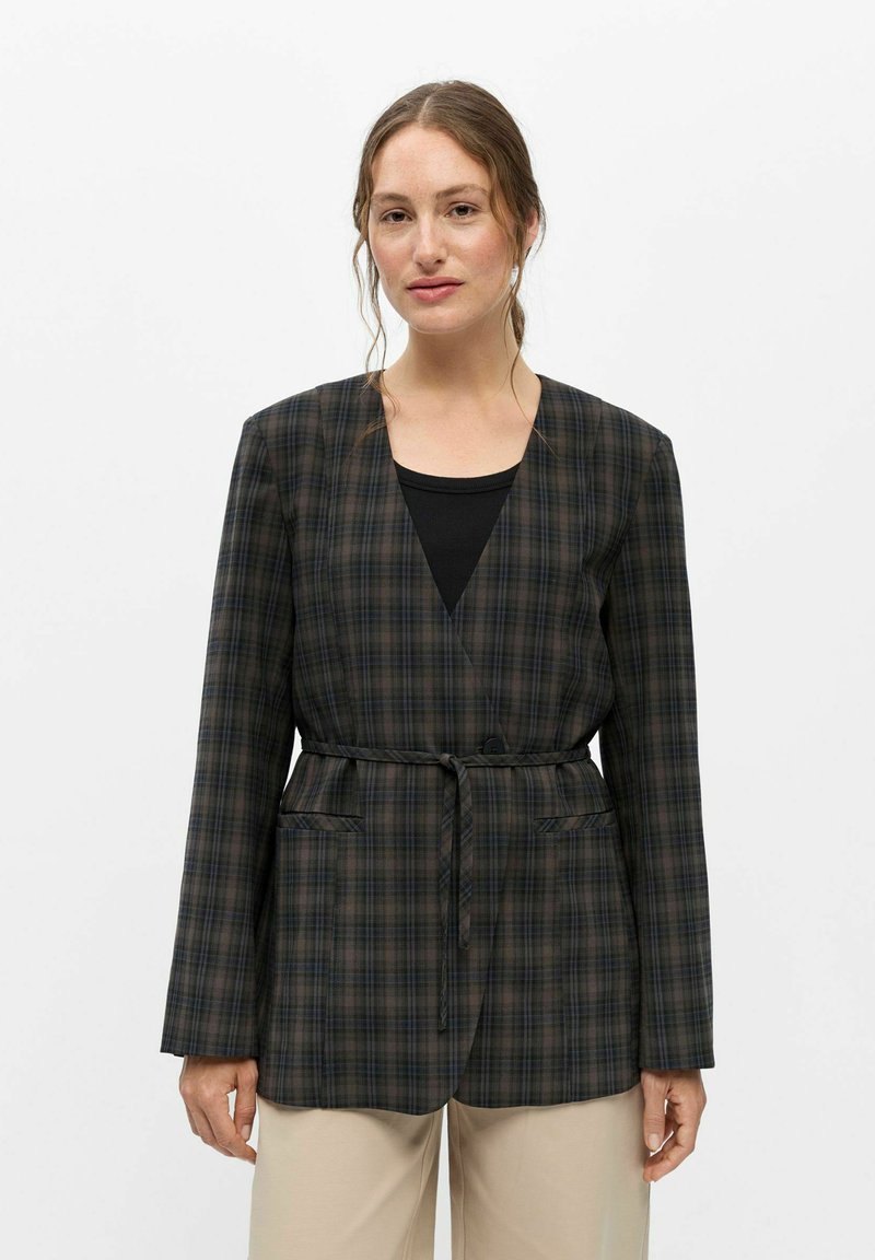 Femme à la peau claire et aux cheveux bruns porte un blazer ceinturé à carreaux foncés sur un haut noir avec un pantalon beige, debout devant un fond blanc.