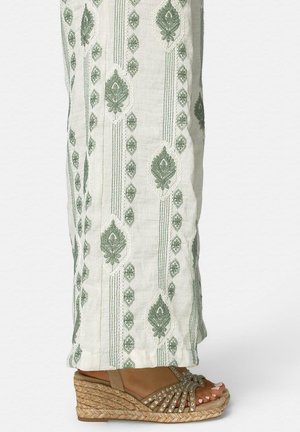 Piernas con pantalones blancos con bordado verde combinados con sandalias cuña color beige con tiras con tachuelas y suelas trenzadas.