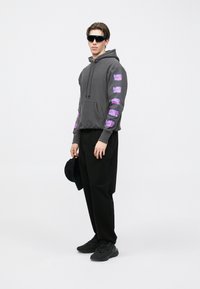 Grauer Kapuzenpullover mit lila grafischen Designs auf den Ärmeln, kombiniert mit schwarzen Hosen und schwarzen Turnschuhen. Trägt eine Sonnenbrille und hält einen Hut in der Hand.