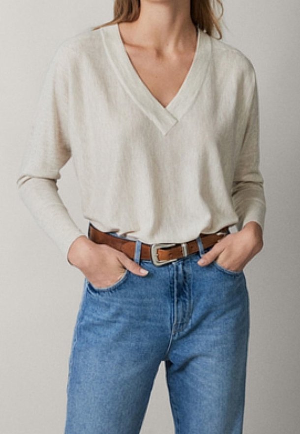 Femme portant un chemisier crème à col en V et manches longues, rentré dans un jean bleu taille haute avec une ceinture en cuir marron, les mains dans les poches.