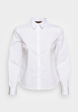 Chemise blanche pour femme à manches longues, boutonnée, avec col pointu et taille cintrée, présentée sur un fond clair uni.