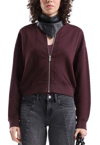 Vrouw in een bordeauxrode kabelgebreide ziptrui, een zwart leren neksjaal en vervaagde zwarte jeans, die een zwart kettingaccessoire vasthoudt.