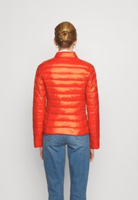 Giacca puffer rossa con finitura lucida, motivo trapuntato e maniche lunghe. Indossata con jeans blu, vista da dietro su uno sfondo semplice.