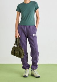 Camiseta verde de manga corta, pantalones de chándal morados con logo blanco, bolso de color oliva y zapatillas beige con cordones azules sobre un fondo verde.