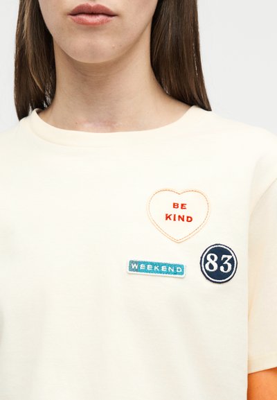 Γυναίκα που φοράει κρεμ πουκάμισο με τρεις μπαλώματα: πορτοκαλί καρδιά με το σύνθημα "ΝΑ ΕΙΣΑΙ ΚΑΛΟΣ", μπλε ορθογώνιο με τη λέξη "ΣΑΒΒΑΤΟΚΥΡΙΑΚΟ" και ναυτικό μπλε κύκλο με τον αριθμό "83".