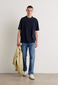 Námořnické bavlněné tričko, volný střih, v kombinaci se světle modrými džíny. Model drží béžovou bundu a na sobě má bílé tenisky s béžovými podrážkami.