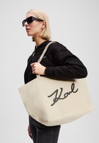 Beige Canvas-Shopper mit schwarzem "Karl"-Logo, ausgestattet mit stabilen Trägern und einem einfachen, geräumigen Design.