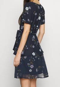 Robe à manches courtes imprimée de fleurs en bleu marine, design en couches avec une taille à volant et un ruban à nouer, présentant des motifs floraux bleus et blancs.