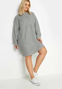 Abito a costine grigio con collo alto e maniche lunghe, caratterizzato da una vestibilità morbida e un orlo sopra il ginocchio. Abbinato a sneakers bianche.