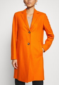 Orange frakk i uldblanding med hakket revers, enkelt knaplukning, to sidelommer og lige snit. Glat tekstur og skræddersyet design.