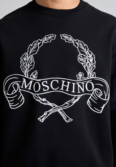 Fekete kapucnis pulóver, fehér hímzett babérlevelekkel és egy "MOSCHINO" felirattal, alatta keresztmintás részletezéssel.