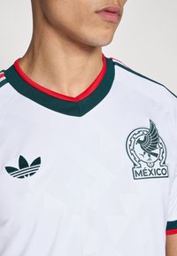Bijeli nogometni dres sa zelenim i crvenim naglascima. Ima V-izrez, Adidas logo i grb reprezentacije Meksika. Teksturirani geometrijski uzorak.