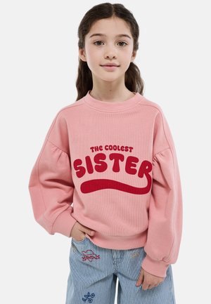 Jeune fille aux longs cheveux foncés portant un sweat-shirt rose sur lequel est inscrit « La sœur la plus cool » et un pantalon bleu à rayures avec de petites broderies.