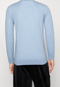 Un pull long à manches longues bleu clair porté par une personne de dos, associé à un pantalon noir en velours ample, le tout sur un fond uni.