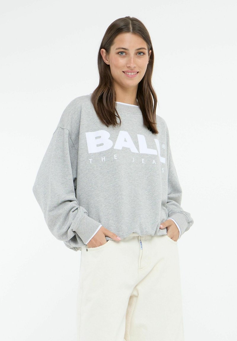 Felpa grigia con testo bianco in grassetto "BALL THE JEANS", vestibilità rilassata, spalle scese e polsini a coste, abbinata a pantaloni chiari.