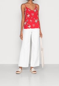 Röd blommig linne med tunna axelremmar, ihop med vita vida byxor och beigea slip-on sandaler. En neutral tote-väska bärs.