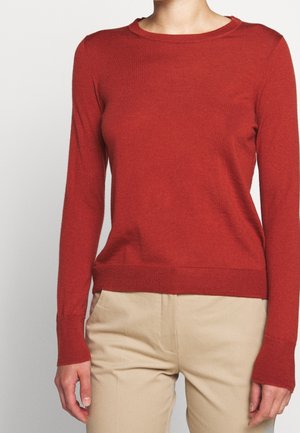 Personne portant un pull rouge ajusté à manches longues et un pantalon beige, debout devant un fond clair uni.