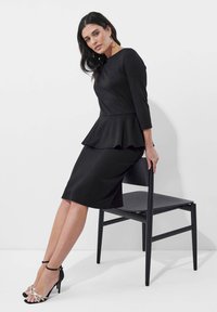 Schwarzes Peplum-Kleid mit dreiviertellangen Ärmeln, tailliertem Oberteil und knielangem Rock. Tragen mit schwarzen und metallischen High-Heels.