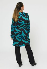 Langærmet kjole i sort med et turkis abstrakt mønster, der har en snøret talje og en flydende silhuet. Kombineret med sorte bukser.