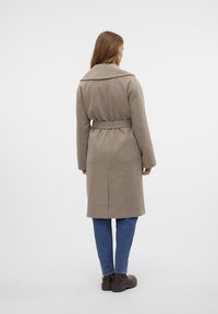 Cappotto in misto lana color sabbia con vita cintata, colletto ampio e spacco posteriore, abbinato a jeans blu e scarpe marroni scure.
