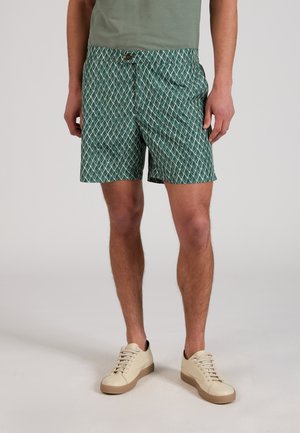 Mężczyzna w zielonych, wzorzystych krótkich spodenkach i beżowych tenisówkach, stojący na jasnym, jednolitym tle.