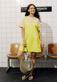 Robe salopette en denim jaune avec un col rond et une poche latérale, associée à un t-shirt beige. Tient un sac hobo en tissu gris et porte des tongs.