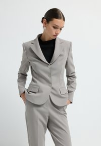 Femme les mains dans les poches, portant un costume ajusté gris clair et un haut noir, regardant vers le bas sur fond blanc uni.
