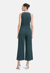 Salopette verde con design senza maniche, corpo aderente, pantaloni larghi, chiusura posteriore con zip e due tasche posteriori. Indossata con tacchi argentati.