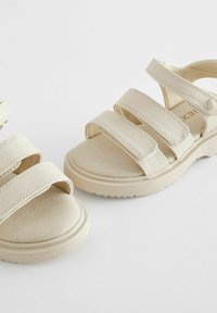 Beiga sandaler med tre justerbara kardborreband, en mjuk mockaliknande textur och en dämpad sula med ett räfflat mönster.