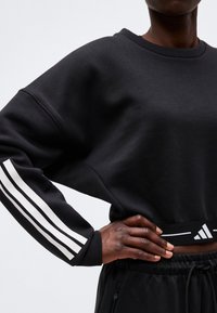 Personne portant un sweat court noir avec des bandes blanches Adidas sur la manche et un logo près de la taille, main reposant sur la hanche.