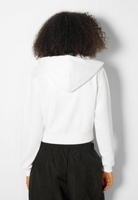 Persona con capelli ricci che indossa un hoodie bianco corto e pantaloni neri con vita elastica, di spalle contro uno sfondo bianco uniforme.