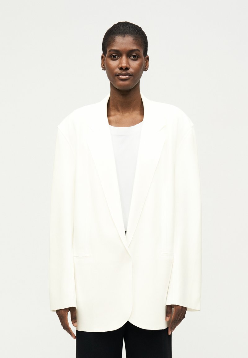 Γυναίκα με κοντά μαλλιά που φοράει ένα φαρδύ λευκό σακάκι και λευκό πουκάμισο σε μια απλή ανοιχτόχρωμη φόντο.