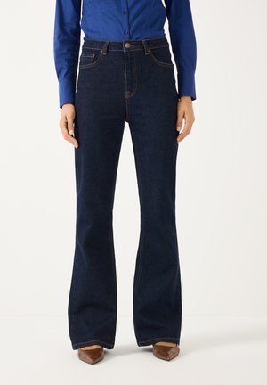 Person trägt dunkelblaue ausgestellte Jeans, blaues Hemd mit Knöpfen und braune spitz zulaufende Schuhe und steht vor einem einfarbigen Hintergrund.