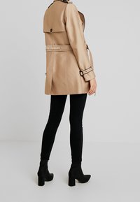 Beżowy, dopasowany płaszcz trencz z paskiem, guziki na ramionach i plecach, noszony na czarnych legginsach i botkach. Gładka faktura.