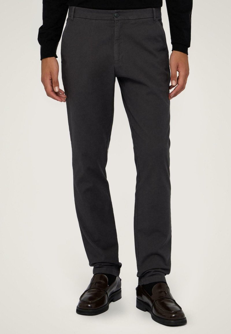 Tmavě šedé kalhoty slim fit, vyrobené z lehkého materiálu. Mají zapínání na knoflík a boční kapsy, nošeny s tmavými mokasíny.