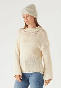 Romige gebreide trui met brede mouwen en een geribde kraag. Draag met lichtblauwe jeans. Beige geribde beanie met een logo patch.