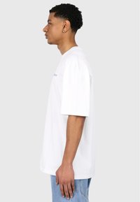 White oversized t-shirt with short sleeves, featuring a crew neckline and embroidered logo in blue on the front. Paired with light blue jeans.