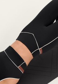 Crne legging hlače s visokim strukom, bijelim obrubima i smeđim detaljem. Karakteriziraju ih elegantan dizajn i glatka tekstura, prikazane na podlozi za jogu.